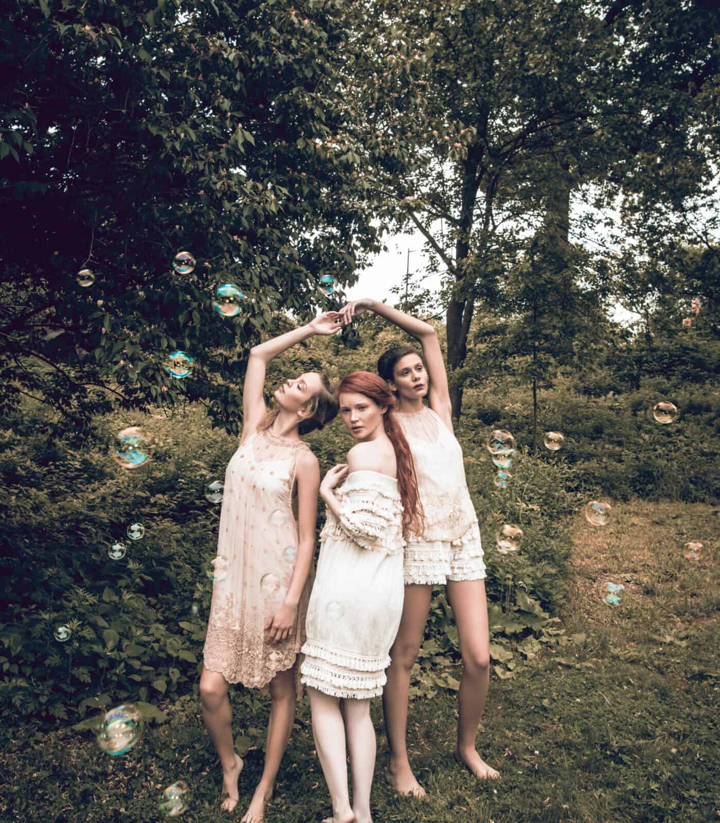 Women in white dresses standing in a lush green outdoor environment with trees and grass.