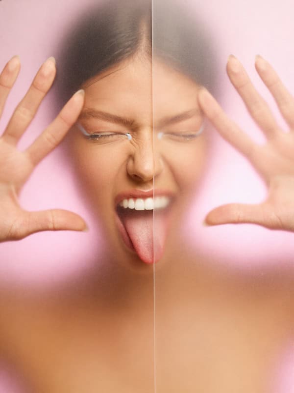 Playful close-up beauty portrait with hands raised and face partially blurred behind frosted pink glass.
