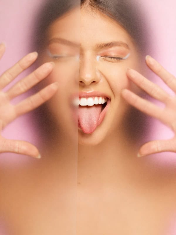 Playful beauty portrait of a model laughing behind frosted pink panels with hands near her face.