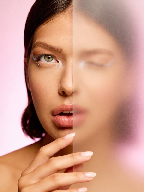 Close-up editorial beauty portrait with one eye visible and part of the face blurred through frosted glass.