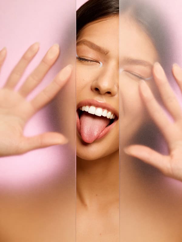 Playful beauty portrait of a model smiling with hands raised behind two frosted glass panels.