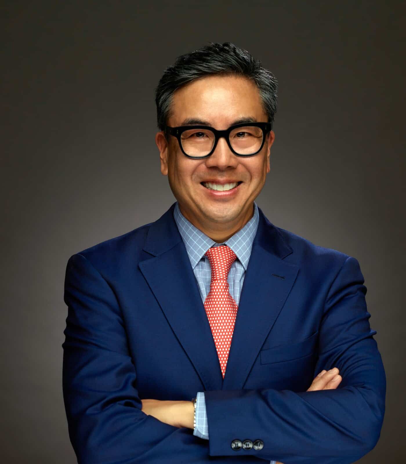 Confident professional man in formal suit with glasses smiling, arms crossed.