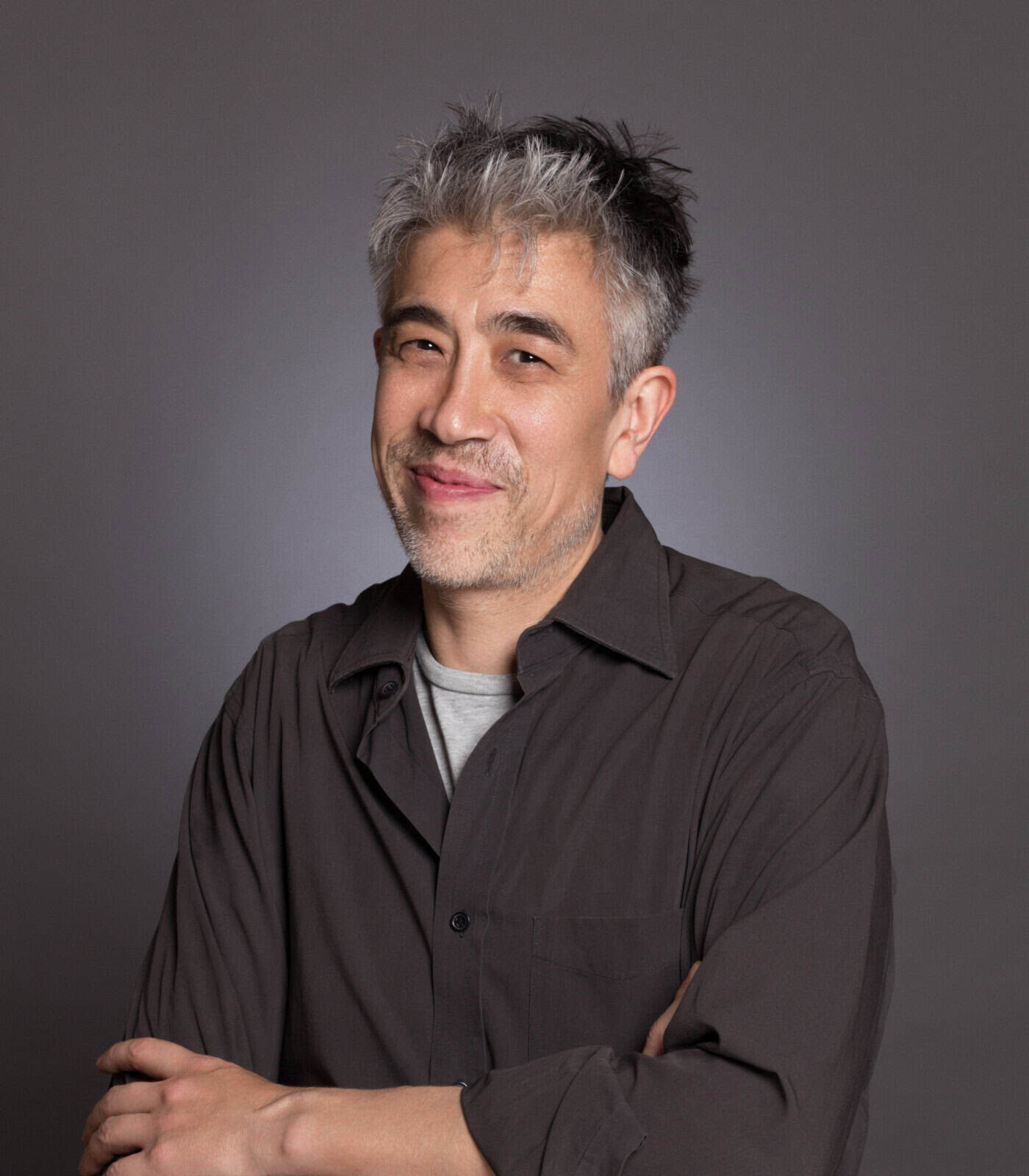Confident middle-aged man with gray hair smiling in a black shirt against gray background.