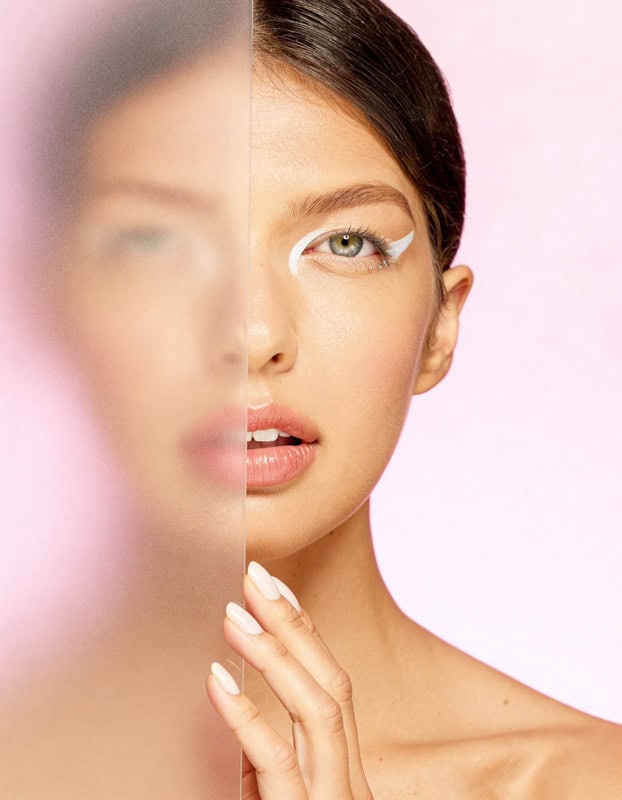 Editorial beauty portrait of a model looking through a vertical frosted glass panel against a pink background.