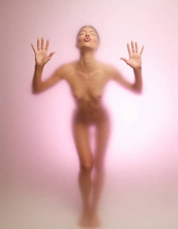 Full-body implied nude beauty portrait photographed through frosted pink glass with hands pressed forward.